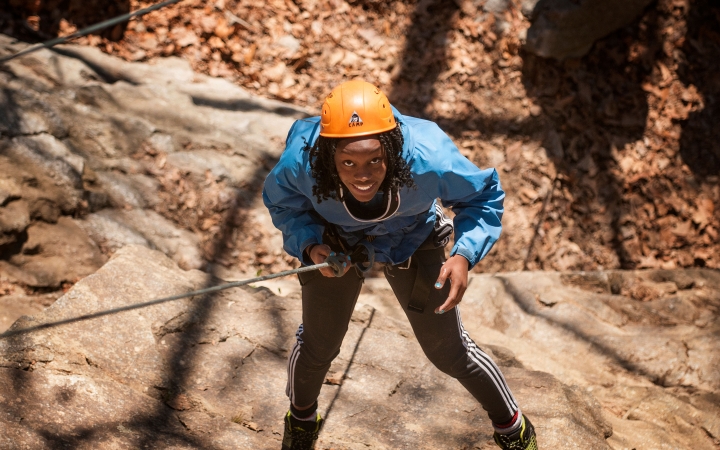 A young person wearing safety gear is secured by ropes as they look up toward the camera and smile while rock climbing. 